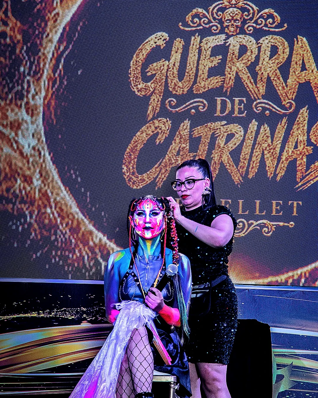 Artista maquillando en vivo a modelo de Catrina neón frente a la pantalla oficial de Guerra de Catrinas de Bellet